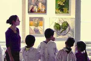 view of several children and a teacher from the back viewing an art exhibit while wearing smocks
