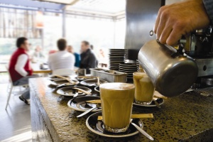 close up image of a cappucino in a mug on a coffee bar with patrons in the background